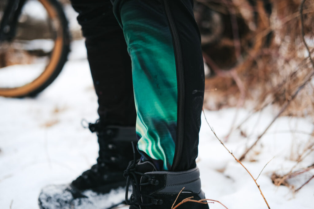 A close up shot of a person leg wearing Borah's OTW XC Pant legs detailing the full length zipper and custom graphics. Snowy landscape in the background.
